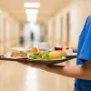 Eine Person in blauer Krankenhauskleidung trägt auf einem Tablett verschiedene Speisen und Getränke durch einen Krankenhausflur. Zu sehen sind unter anderem Brötchen, Salat, Dessert und eine Tasse.
