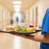 Eine Person in blauer Krankenhauskleidung trägt auf einem Tablett verschiedene Speisen und Getränke durch einen Krankenhausflur. Zu sehen sind unter anderem Brötchen, Salat, Dessert und eine Tasse.