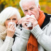 Eine ältere Frau und ein älterer Mann stehen vor herbstlich verfärbten Bäumen und schnäuzen sich in Taschentücher