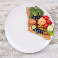 Gabel und Löffel liegen angeordnet wie Zeiger einer Uhr auf einem Teller, dazwischen liegen Lebensmittel