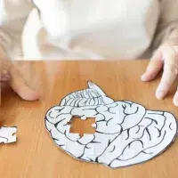 Symbolbild: Eine Person sitzt vor einem Gehirn-Puzzle mit fehlendem Teil auf einem Holztisch