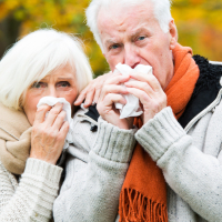 Eine ältere Frau und ein älterer Mann stehen vor herbstlich verfärbten Bäumen und schnäuzen sich in Taschentücher