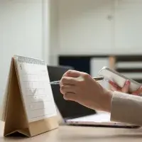 Nahaufnahme der Hand einer Frau, die einen Stift hält und im Begriff ist, etwas in einen Tischkalender einzutragen.