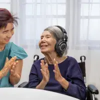 Eine junge und eine ältere Frau sitzen zueinandergewandt an einem Tisch. Die ältere Frau hört Musik über Kopfhörer. Beide klatschen in ihre Hände.