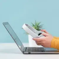 Laptop steht auf einem Tisch, darüber halten zwei Hände einen Kopfhörer fest