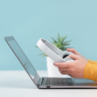 Laptop steht auf einem Tisch, darüber halten zwei Hände einen Kopfhörer fest